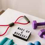From above composition of dumbbells and massage double ball and tape and tubular expanders surrounding light box with wake up and workout words placed on white surface of table